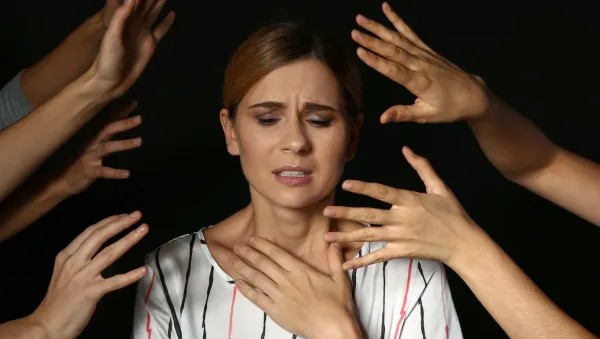 Woman looks anxious as multiple hands reach toward her neck and shoulders against a dark background.