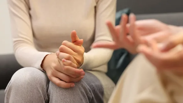 Person in a beige sweater sits with hands clasped on their lap while another person gestures with open hands during a counseling session.