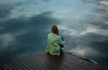 Person sits with back to camera on a wooden dock, looking out over calm water reflecting the sky and clouds.