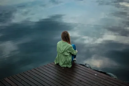 Person sits with back to camera on a wooden dock, looking out over calm water reflecting the sky and clouds.