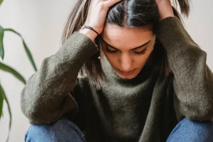 Young woman sits with head in hands, looking down in distress, wearing an olive-green sweater and jeans in a neutral room.