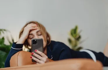 Woman lying on a brown couch, propping her head with one hand while looking at a smartphone in the other hand.