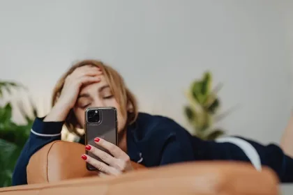 Woman lying on a brown couch, propping her head with one hand while looking at a smartphone in the other hand.