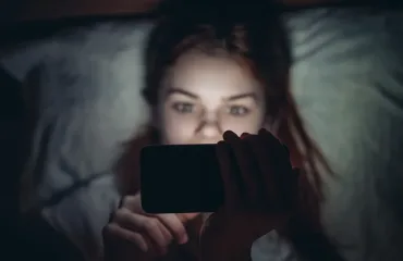Person lying in bed at night, holding a smartphone with the screen glowing in their face.