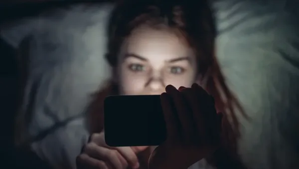 Person lying in bed at night, holding a smartphone with the screen glowing in their face.
