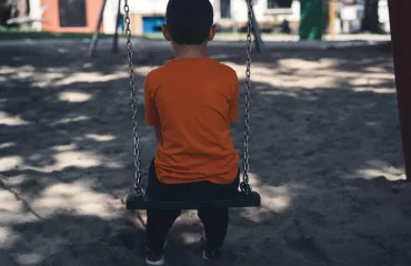 Child sitting on a swing facing away in a sunny playground, wearing an orange shirt.