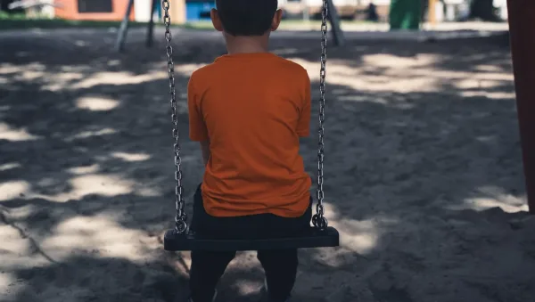 Childhood Trauma and Adult Relationships How Therapy Helps Child sitting on a swing facing away in a sunny playground, wearing an orange shirt.