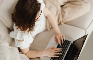 How Online Therapy Makes Mental Health Support More Accessible Person with brown hair using a laptop on a beige sofa, seen from above, wearing a light top and glasses
