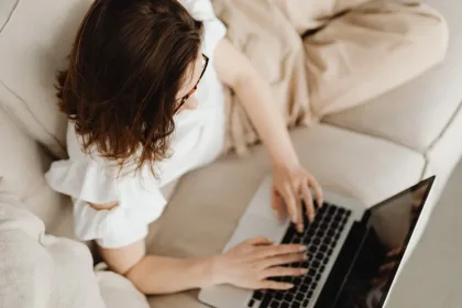 Person with brown hair using a laptop on a beige sofa, seen from above, wearing a light top and glasses