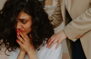 A woman with curly hair covers her mouth in distress as another person places a comforting hand on her shoulder.