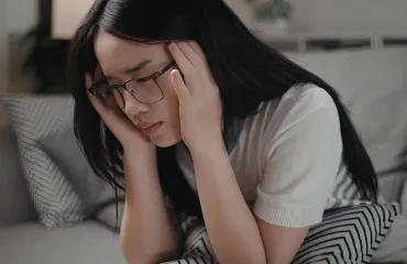Woman wearing glasses sits on a couch, holding her head in her hands and looking stressed or overwhelmed.