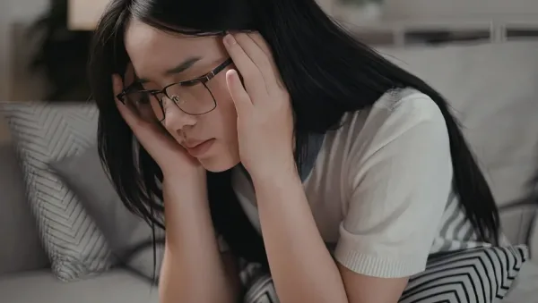 Is It Stress or Anxiety How to Recognize the Difference Woman wearing glasses sits on a couch, holding her head in her hands and looking stressed or overwhelmed.