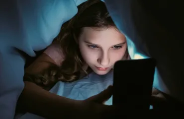 Girl lying in bed under a blue blanket, glow from a smartphone lighting her face as she looks at the screen