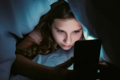 Girl lying in bed under a blue blanket, glow from a smartphone lighting her face as she looks at the screen
