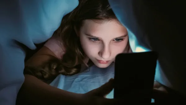 Screen Addiction in Teens How Therapy Supports Families Girl lying in bed under a blue blanket, glow from a smartphone lighting her face as she looks at the screen