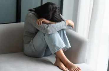 Person sits curled on a light gray couch, arms resting on their knees with head down, conveying distress or sadness.