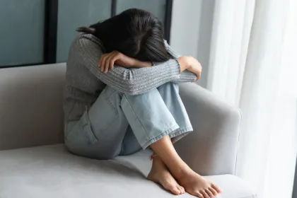 Person sits curled on a light gray couch, arms resting on their knees with head down, conveying distress or sadness.