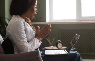 Woman sits on a sofa with hands in prayer, looking at an open laptop on her lap.