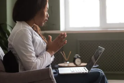 Woman sits on a sofa with hands in prayer, looking at an open laptop on her lap.