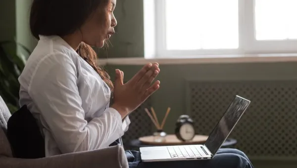 Woman sits on a sofa with hands in prayer, looking at an open laptop on her lap.
