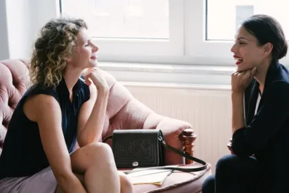 Two women sit on a pink couch by a window, smiling and talking in a bright room.