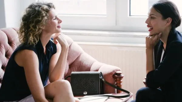 Two women sit on a pink couch by a window, smiling and talking in a bright room.