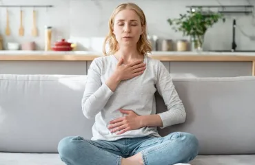 Woman sits cross-legged on a sofa with eyes closed, hands on chest and stomach, practicing deep breathing in a bright kitchen.