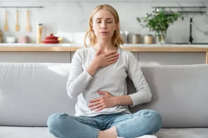 Woman sits cross-legged on a sofa with eyes closed, hands on chest and stomach, practicing deep breathing in a bright kitchen.