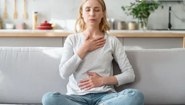 Woman sits cross-legged on a sofa with eyes closed, hands on chest and stomach, practicing deep breathing in a bright kitchen.