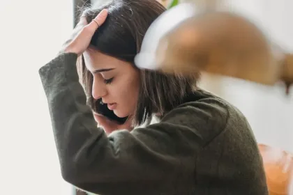 Young woman with dark hair sits indoors, hand on her forehead and looking down in a stressed, contemplative pose.