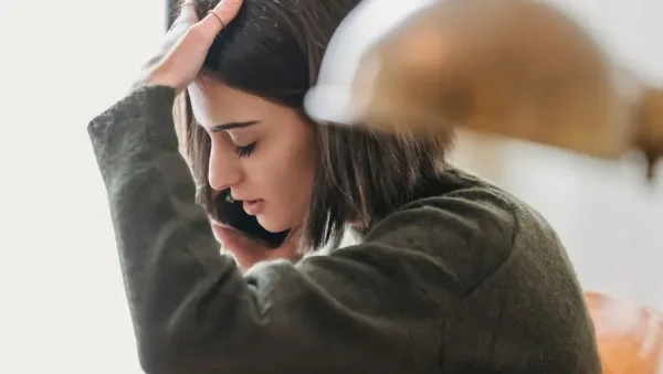 Young woman with dark hair sits indoors, hand on her forehead and looking down in a stressed, contemplative pose.