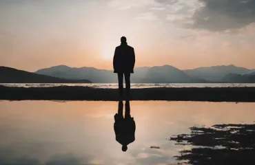 Silhouette of a person standing on a shore at sunset, with mountains in the background and a reflection in the calm water.