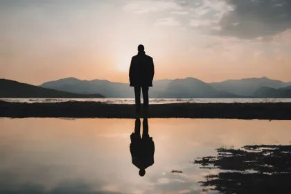 Silhouette of a person standing on a shore at sunset, with mountains in the background and a reflection in the calm water.