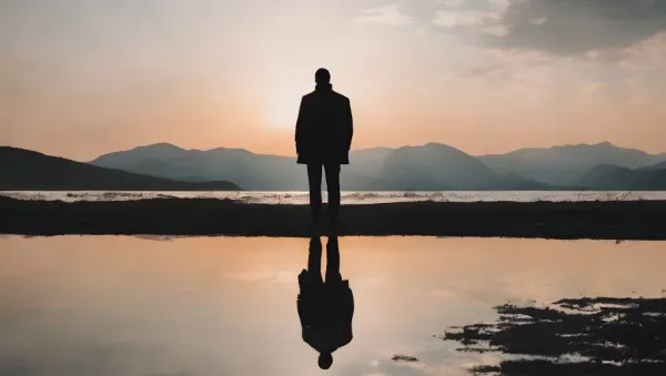 Silhouette of a person standing on a shore at sunset, with mountains in the background and a reflection in the calm water.