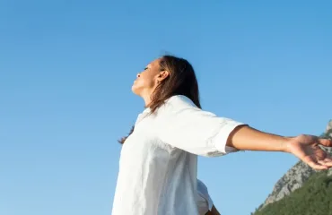 Woman in a white shirt stands with arms outstretched under a clear blue sky, mountains in the background.