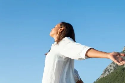 Woman in a white shirt stands with arms outstretched under a clear blue sky, mountains in the background.