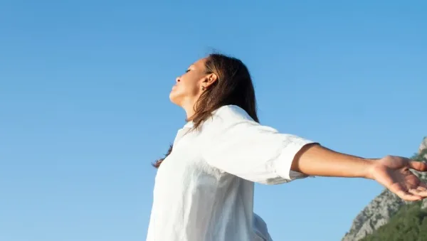 Woman in a white shirt stands with arms outstretched under a clear blue sky, mountains in the background.