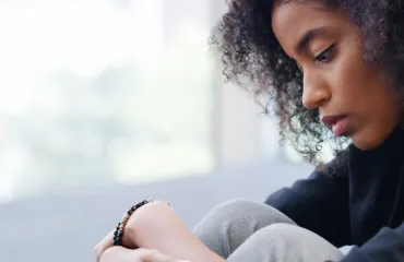 Emotional Flashbacks What They Are & How to Cope Young woman with curly hair sits with knees drawn up, looking down in thought on a bright indoor setting.