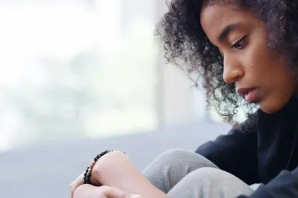 Young woman with curly hair sits with knees drawn up, looking down in thought on a bright indoor setting.