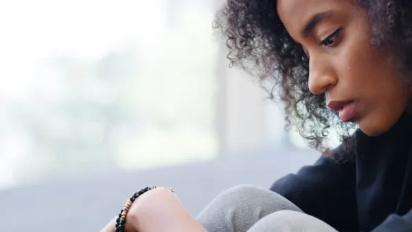 Young woman with curly hair sits with knees drawn up, looking down in thought on a bright indoor setting.