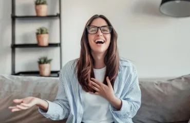 Woman with long brown hair and glasses laughs while seated on a grey sofa at home, hand on chest.
