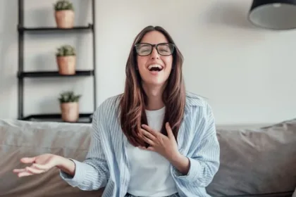 Woman with long brown hair and glasses laughs while seated on a grey sofa at home, hand on chest.