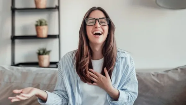 Woman with long brown hair and glasses laughs while seated on a grey sofa at home, hand on chest.