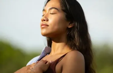 Woman outdoors with eyes closed, basking in sunlight, hands holding a light jacket over her shoulders.