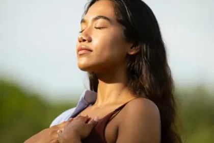 Woman outdoors with eyes closed, basking in sunlight, hands holding a light jacket over her shoulders.