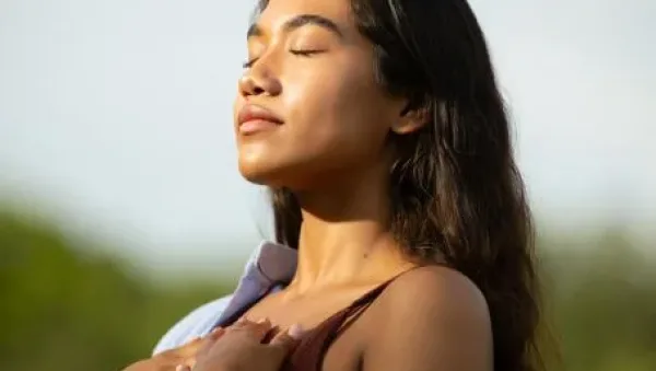 Woman outdoors with eyes closed, basking in sunlight, hands holding a light jacket over her shoulders.