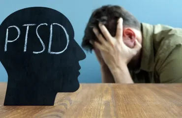 Silhouette of a head with 'PTSD' written inside, facing a man who holds his head in distress at a wooden table.