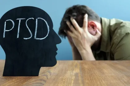 Silhouette of a head with 'PTSD' written inside, facing a man who holds his head in distress at a wooden table.