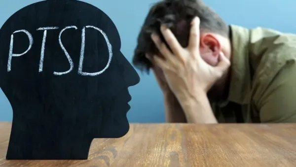 Silhouette of a head with 'PTSD' written inside, facing a man who holds his head in distress at a wooden table.